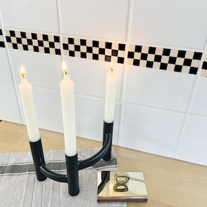 Op de afbeelding: Een zwarte metalen kandelaar met drie witte kaarsen, elk met een gele vlam. De houder staat op een grijs-wit gestreepte doek, met een goudkleurige lucifersdoos in de buurt. De achtergrond heeft witte tegels met een zwart-wit geblokte rand.