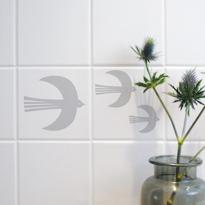 Puede incluir: Tres siluetas de pájaros grises se adhieren a azulejos blancos. Los pájaros vuelan en diferentes direcciones. Un jarrón con una planta verde y flores de cardo azul se encuentra en la esquina de la imagen.