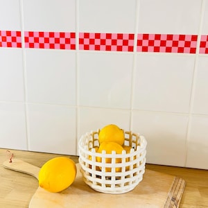 May include: White ceramic bowl with a grid pattern, filled with yellow lemons, sitting on a wooden surface. A pink and red checkered border is visible on the white tiled wall behind the bowl.