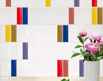 May include: A wall of white square tiles featuring vertical stripes in red, yellow, purple, blue, and brown. A white textured vase holds pink peony flowers, and black metal scissors lie on a light wood surface with a few green leaves.