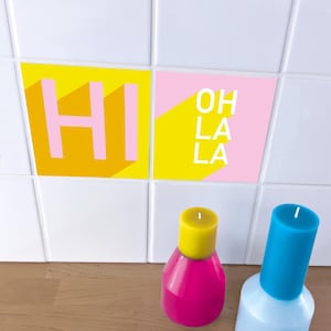 Puede incluir: Dos velas y dos baldosas cuadradas con texto. Una baldosa tiene la palabra "HI" en rosa sobre un fondo amarillo. La otra baldosa tiene las palabras "OH LA LA" en blanco sobre un fondo rosa y amarillo. Una vela es rosa con la parte superior amarilla, y la otra es azul claro con la parte superior azul.