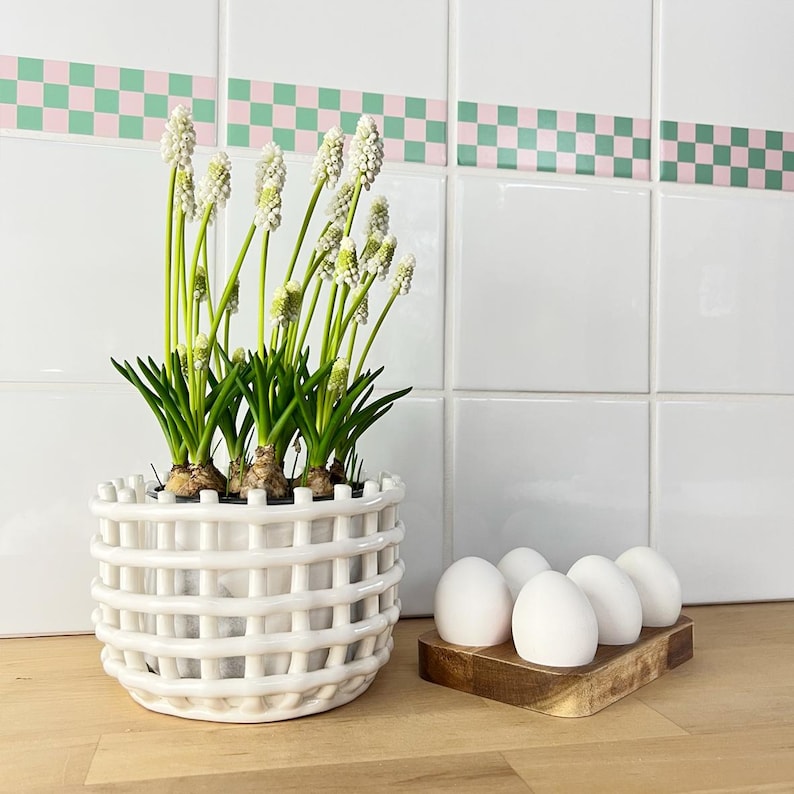May include: A white ceramic planter with a lattice design holds white flowers with green stems and leaves. Six white eggs sit in a wooden egg holder on a wooden surface. The background features white tiles with a pink and green checkered border.