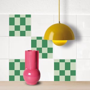 Può includere: Un vaso in ceramica rosa con corpo rotondo e collo alto e stretto si trova su una superficie di legno di fronte a una parete piastrellata bianca con piastrelle a scacchi verdi e bianche. Una lampada a sospensione gialla pende dal soffitto sopra il vaso.