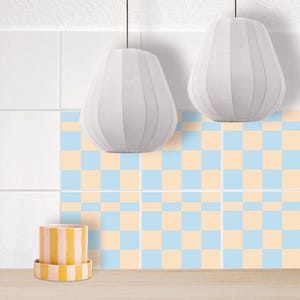 Puede incluir: Dos lámparas colgantes blancas en forma de pera cuelgan sobre una pared con un patrón de tablero de ajedrez de cuadrados azul claro y melocotón. Un macetero amarillo a rayas se encuentra en la encimera debajo de la pared.