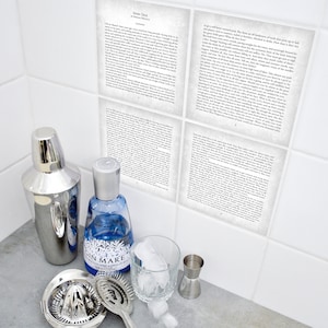 Puede incluir: Una coctelera de plata, una botella de ginebra de vidrio transparente con una etiqueta azul que dice "Gin Mare", un medidor de plata, un exprimidor de cítricos, un colador de cóctel de plata y un vaso con cubitos de hielo. El fondo es una pared de azulejos blancos con cuatro azulejos que tienen texto impreso en ellos. El texto en los azulejos es del libro "More Deck" de Herman Melville.