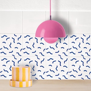 Puede incluir: Una lámpara colgante rosa con un techo blanco, una pared de azulejos blancos con un patrón geométrico azul y una maceta rayada amarilla y rosa sobre una encimera blanca.