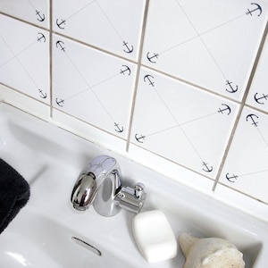 Peut inclure: Un lavabo de salle de bain blanc avec un robinet chromé, un savon et une coquille de mer. Le mur derrière le lavabo est carrelé de carreaux blancs et décoré de petits symboles d'ancres.