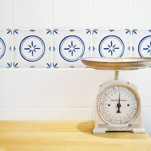 Puede incluir: Una báscula de cocina blanca con un salpicadero de azulejos azules y blancos. La báscula tiene un dial redondo con números negros y el texto "Alexanderwerk" impreso en él.