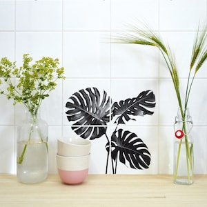 Puede incluir: Calcomanías de hojas de monstera en blanco y negro sobre azulejos blancos. Las calcomanías están dispuestas en un patrón de tres hojas.