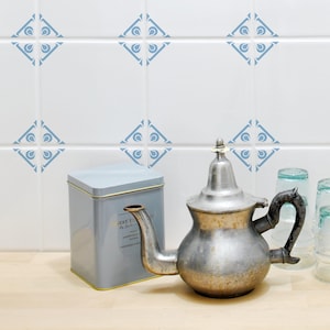 Puede incluir: Una tetera plateada vintage con un pico curvo y un asa negra se encuentra sobre una superficie de madera. También hay una lata de té de metal gris y dos vasos transparentes. El fondo presenta azulejos blancos con un patrón geométrico azul.