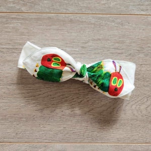 May include: A white headband featuring a green and red caterpillar design. The caterpillar has two large eyes and a red body with green stripes.