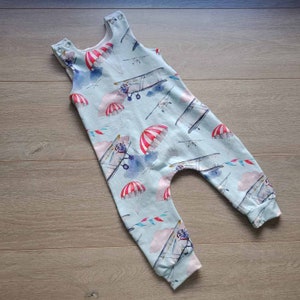 Puede incluir: Un pelele de bebé azul claro con estampado de aviones y paracaídas. El pelele es sin mangas y tiene un cierre de presión en la entrepierna.