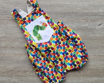 May include: A colorful baby romper with a pattern of multi-colored polka dots. The romper features a white pocket with an illustration of a green caterpillar. The straps and leg openings are elasticized. The fabric is a bright mix of colors.