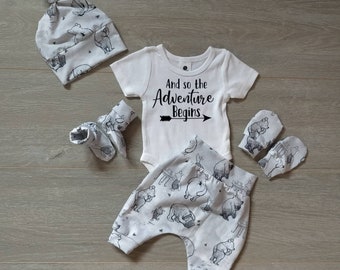 May include: A white baby outfit set with a white onesie that says "And so the Adventure Begins" with a black arrow, white pants with a black bear print, a white hat with a black bear print, and white mittens with a black bear print.