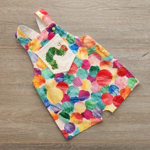 May include: A brightly coloured baby romper featuring a pattern of large, overlapping circles in various colours. The romper has a small pocket with an embroidered illustration of a green caterpillar. White straps with green buttons.