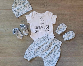 May include: A baby outfit set with a white bodysuit with the text "Hi I'm New Here" and an arrow, blue and white floral print pants, a matching hat, and two pairs of mittens.