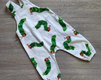 May include: White baby overalls with a green and red caterpillar print. The overalls have a button closure and adjustable straps.
