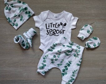 May include: A baby clothing set featuring a white onesie with "Little Sprout" text, matching pants, a knotted hat, socks, and mittens. The set has a green eucalyptus leaf pattern on a white background. The items are arranged on a wooden surface.
