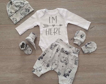 May include: A baby outfit set with a white long-sleeved bodysuit with the text "I'm Here" and an arrow with a heart, gray and white patterned pants, a gray and white patterned hat, and a pair of gray and white patterned booties.