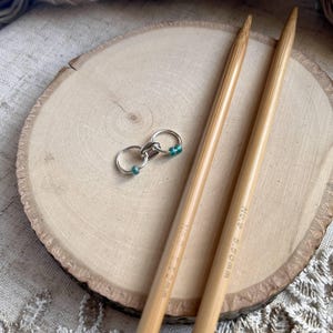 May include: Two light-colored bamboo knitting needles with the text 5.50mm printed on the surface, accompanied by a small silver-toned metal stitch marker featuring two interconnected rings and two tiny teal-colored beads attached to the metal loops.