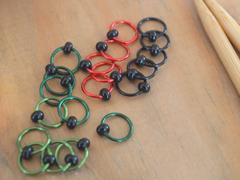 Set of knitting stitch markers on wood background. Rings are lime, green, red and black with black beads.