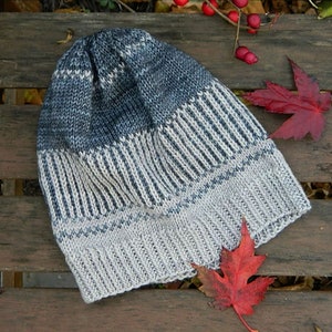 May include: A gray and white knitted beanie hat with a striped pattern. The hat is lying on a wooden surface with red maple leaves.