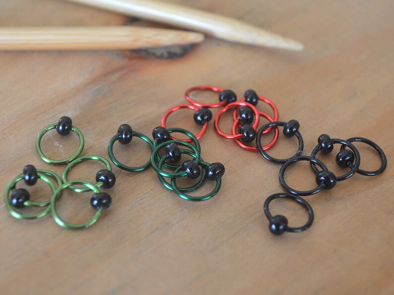 Set of knitting stitch markers on wood background. Rings are lime, green, red and black with black beads.