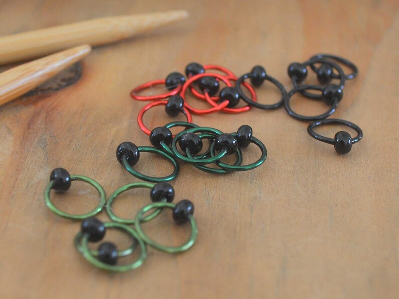 Set of knitting stitch markers on wood background. Rings are lime, green, red and black with black beads.