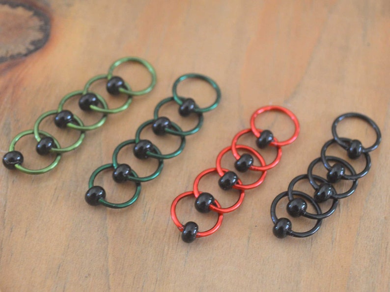 Set of knitting stitch markers on wood background arranged by color. Rings are lime, green, red and black with black beads.