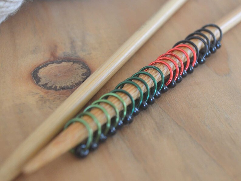 Set of knitting stitch markers on wood background displayed on knitting needle by color. Rings are lime, green, red and black with black beads.