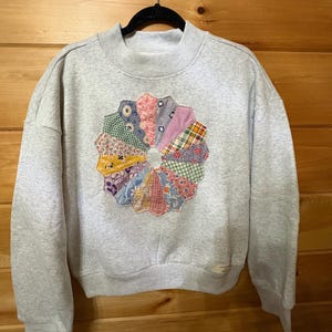 May include: A light gray crewneck sweatshirt featuring a central circular applique made of various colorful patterned fabric segments arranged in a fan-like design. The garment has long sleeves, a ribbed collar, and a small rectangular label at the hem.