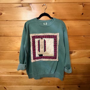 May include: A sage green crewneck sweatshirt featuring a large, rectangular, quilted fabric patch on the front. The patch displays a geometric pattern in cream, black, and magenta, with a central design resembling a stylized boot silhouette.