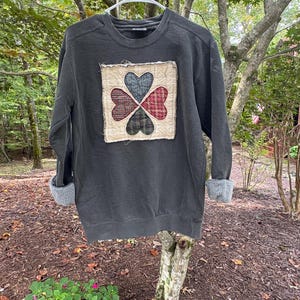 May include: A charcoal gray long-sleeve sweatshirt featuring a square fabric patch on the chest. The patch displays four heart shapes arranged in a clover pattern, crafted from red and dark green plaid fabric with frayed edges.
