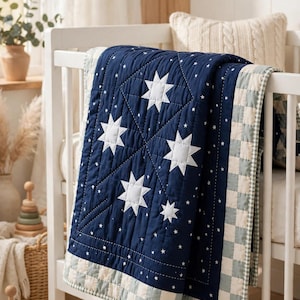 Può includere: Una trapunta blu navy con applicazioni a forma di stella bianca e un motivo a piccole stelle. La trapunta è bordata da un tessuto a scacchi verde chiaro e bianco ed è drappeggiata sul lato di una struttura per culla in legno bianco.