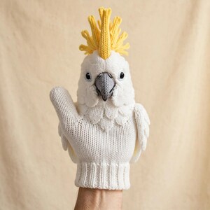 May include: A white knitted mitten shaped like a cockatoo bird. The mitten features a bright yellow crest on top, a grey beak, and black eyes. The body of the mitten is white with textured knit details representing feathers and wings on the sides.