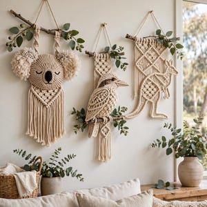 May include: Three macrame wall hangings made of cream-colored cord suspended from wooden branches. The designs feature a koala head with fluffy ears, a kookaburra bird perched on a branch, and a kangaroo silhouette, all accented with green eucalyptus leaves.