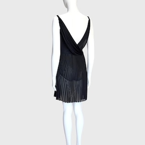 K&ouml;nnte beinhalten: Ein schwarzes Kleid mit tiefem V-Ausschnitt und plissiertem Rock. Das Kleid hat d&uuml;nne Tr&auml;ger und ein drapiertes R&uuml;ckendetail. Der Rock hat ein halbtransparentes Aussehen. Das Kleid wird auf einer Schaufensterpuppe pr&auml;sentiert.