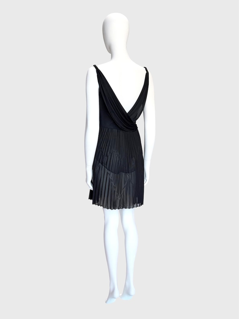 K&ouml;nnte beinhalten: Ein schwarzes Kleid mit tiefem V-Ausschnitt und plissiertem Rock. Das Kleid hat d&uuml;nne Tr&auml;ger und ein drapiertes R&uuml;ckendetail. Der Rock hat ein halbtransparentes Aussehen. Das Kleid wird auf einer Schaufensterpuppe pr&auml;sentiert.
