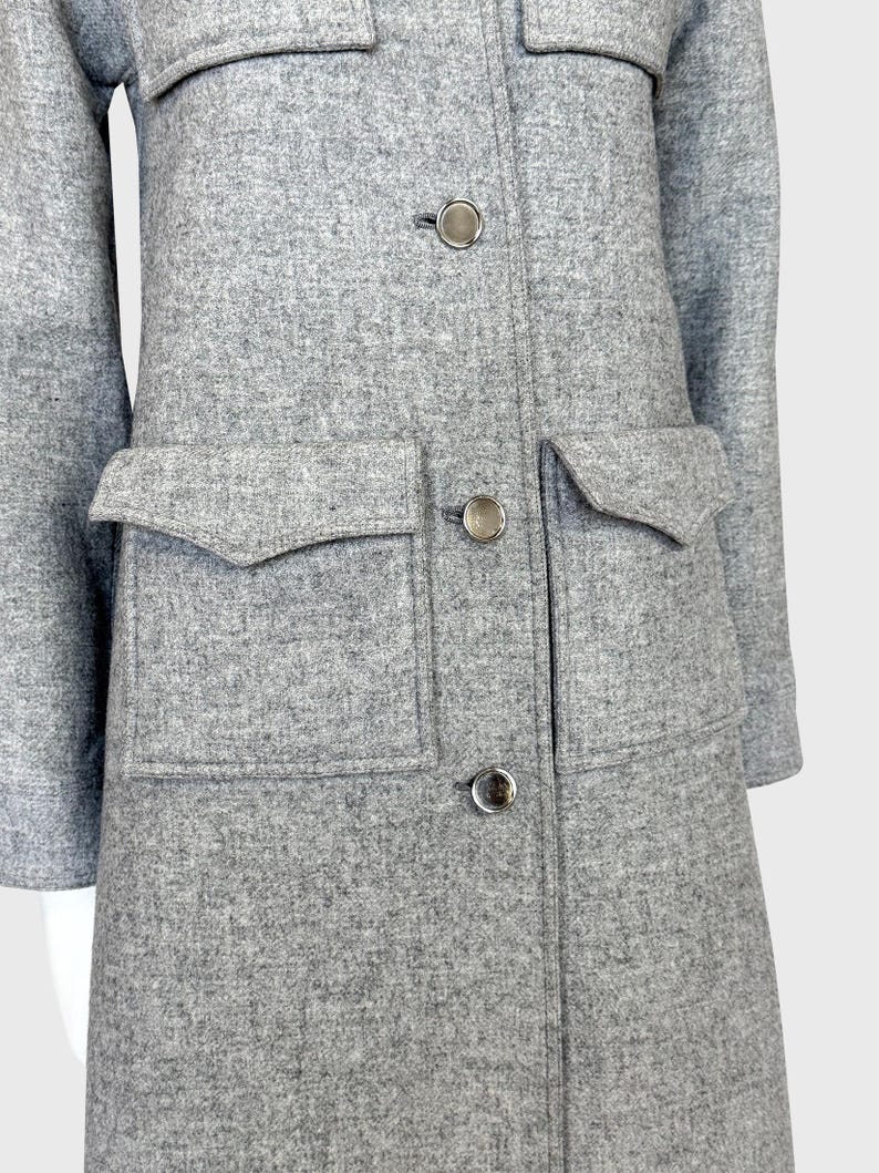 May include: A light gray coat with a textured fabric. The coat features large flap pockets, silver buttons, and a classic collar. The coat's design is simple and elegant, suitable for various occasions.