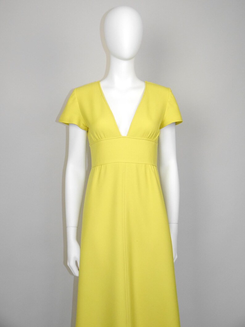 May include: A bright yellow dress with a V-neckline and short sleeves. The dress has a fitted waist and a flowing skirt. The dress is displayed on a mannequin. The dress is a solid color.