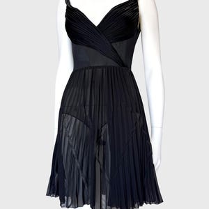 K&ouml;nnte beinhalten: Schwarzes Cocktailkleid auf einer Schaufensterpuppe. Das Kleid hat einen plissierten Rock und ein Oberteil mit V-Ausschnitt. Die Tr&auml;ger sind schmal und das Kleid scheint aus flie&szlig;endem Stoff zu sein.