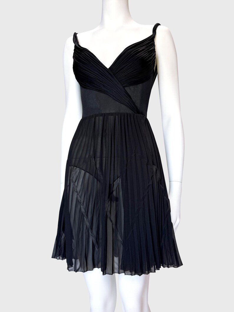 K&ouml;nnte beinhalten: Schwarzes Cocktailkleid auf einer Schaufensterpuppe. Das Kleid hat einen plissierten Rock und ein Oberteil mit V-Ausschnitt. Die Tr&auml;ger sind schmal und das Kleid scheint aus flie&szlig;endem Stoff zu sein.