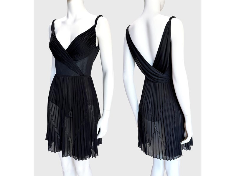 K&ouml;nnte beinhalten: Ein schwarzes Kleid mit einem plissierten Rock und einem V-Ausschnitt-Oberteil. Das Kleid hat d&uuml;nne Tr&auml;ger und einen tiefen R&uuml;cken. Das Oberteil hat ein Wickeldesign, und der Rock ist halbtransparent, wodurch eine flie&szlig;ende Silhouette entsteht. Das Kleid wird auf einer Schaufensterpuppe pr&auml;sentiert.