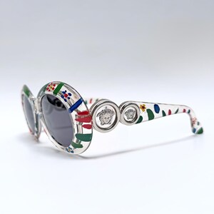 May include: Clear oval sunglasses with a colorful floral pattern on the frames and arms. The sunglasses feature dark lenses and silver Medusa head emblems on the temples. A stylish accessory for eye protection.