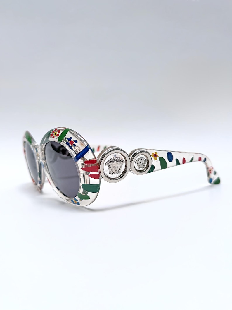 May include: Clear oval sunglasses with a colorful floral pattern on the frames and arms. The sunglasses feature dark lenses and silver Medusa head emblems on the temples. A stylish accessory for eye protection.