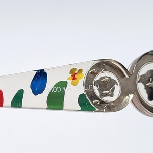 May include: Close-up of a pair of sunglasses with a white arm featuring colorful paint splotches in red, blue, green, and yellow. The silver frame has a Medusa head logo. The text "MOD.418 COL.CD" is printed on the arm.