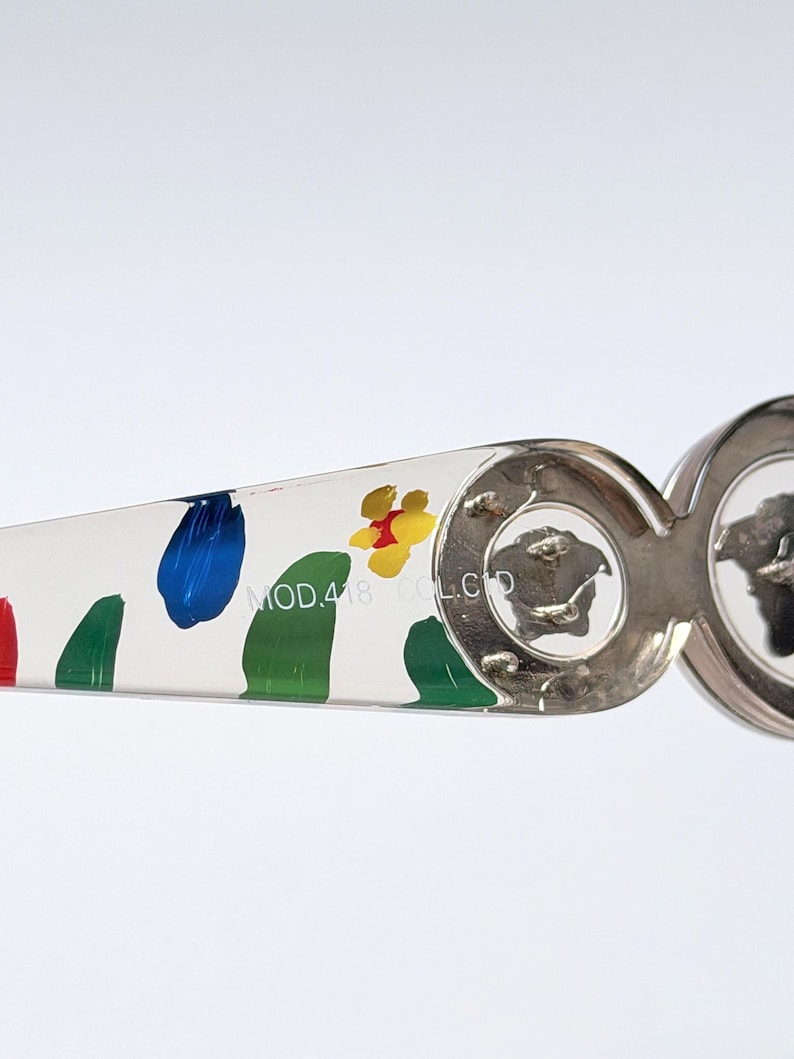 May include: Close-up of a pair of sunglasses with a white arm featuring colorful paint splotches in red, blue, green, and yellow. The silver frame has a Medusa head logo. The text "MOD.418 COL.CD" is printed on the arm.