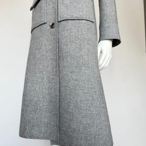 May include: A light gray wool coat with a flared skirt and long sleeves. The coat features two front pockets and gold-colored buttons. The coat is displayed on a mannequin.