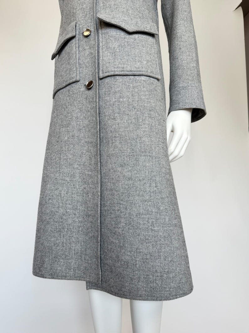 May include: A light gray wool coat with a flared skirt and long sleeves. The coat features two front pockets and gold-colored buttons. The coat is displayed on a mannequin.
