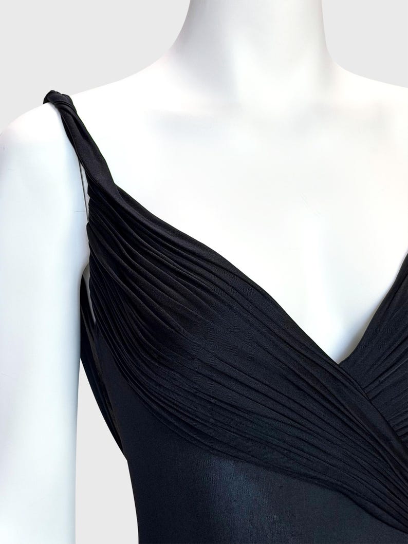 K&ouml;nnte beinhalten: Ein schwarzes Kleid mit drapiertem Oberteil und V-Ausschnitt. Das Kleid hat ein plissiertes Design &uuml;ber der Brust und Tr&auml;ger. Der Stoff scheint ein weiches, flie&szlig;endes Material zu sein.